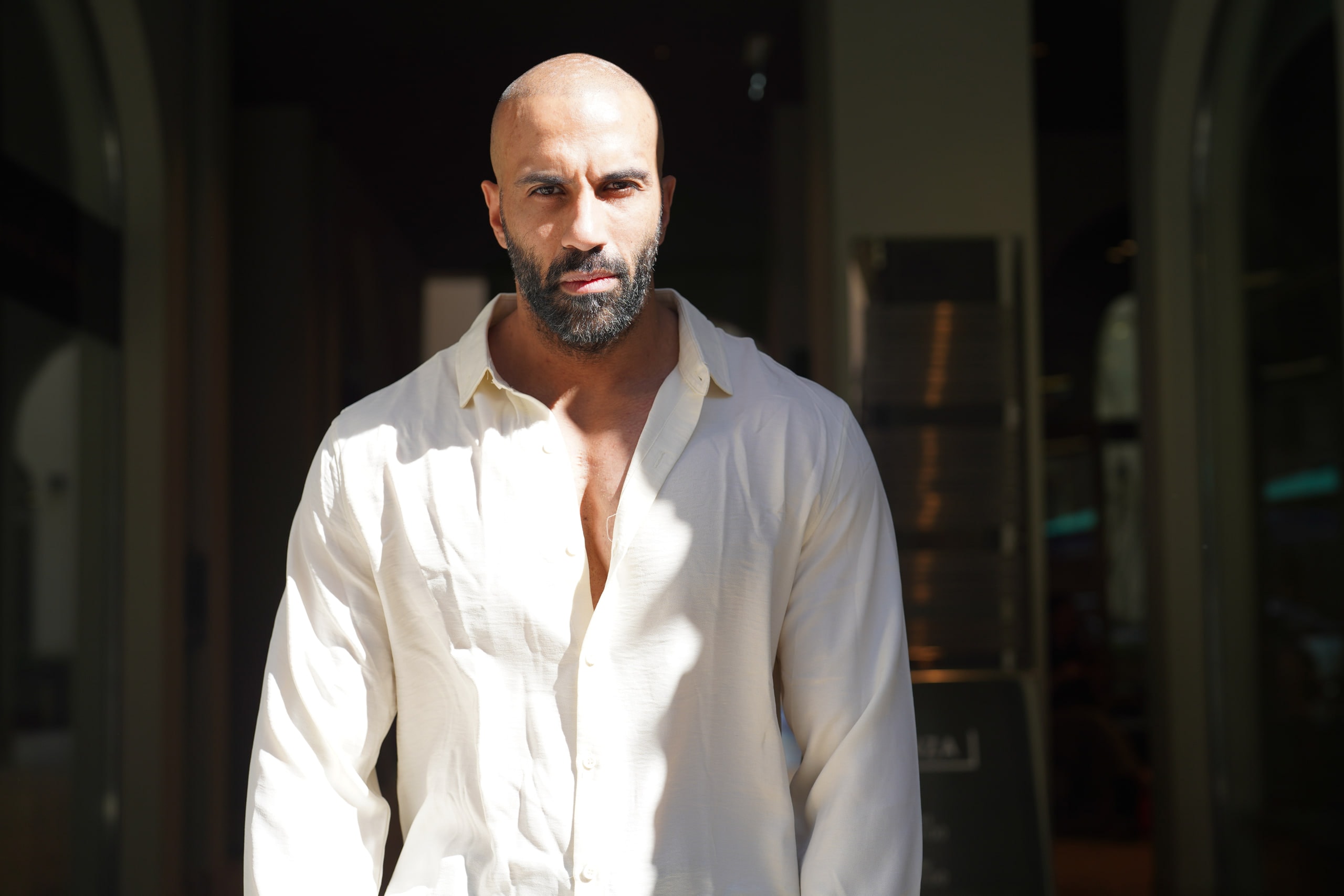 Amer El-Erwadi in an open-collar white shirt, standing in strong sunlight against a dark doorway.