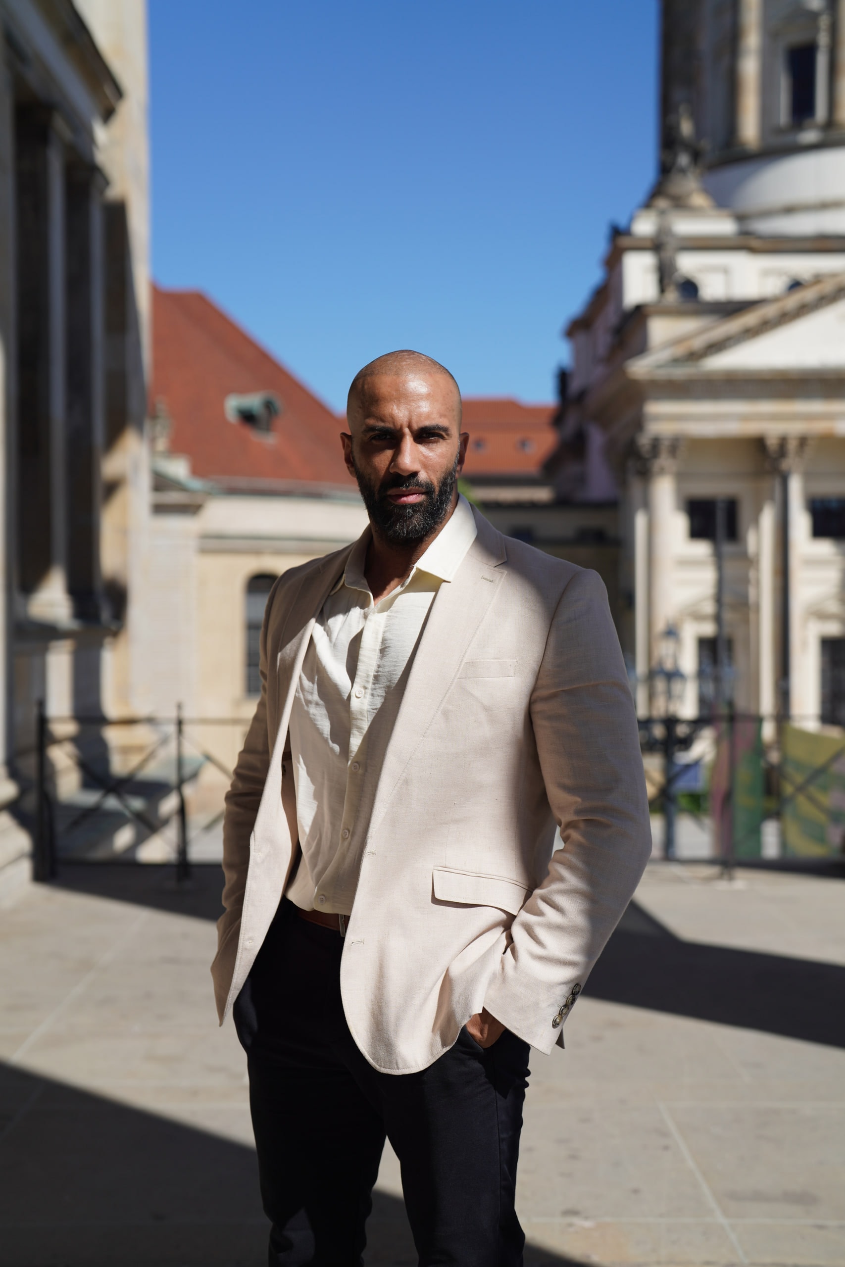 Amer El-Erwadi in a cream jacket with one hand in his pocket, framed by the architecture of Gendarmenmarkt beneath a blue sky.