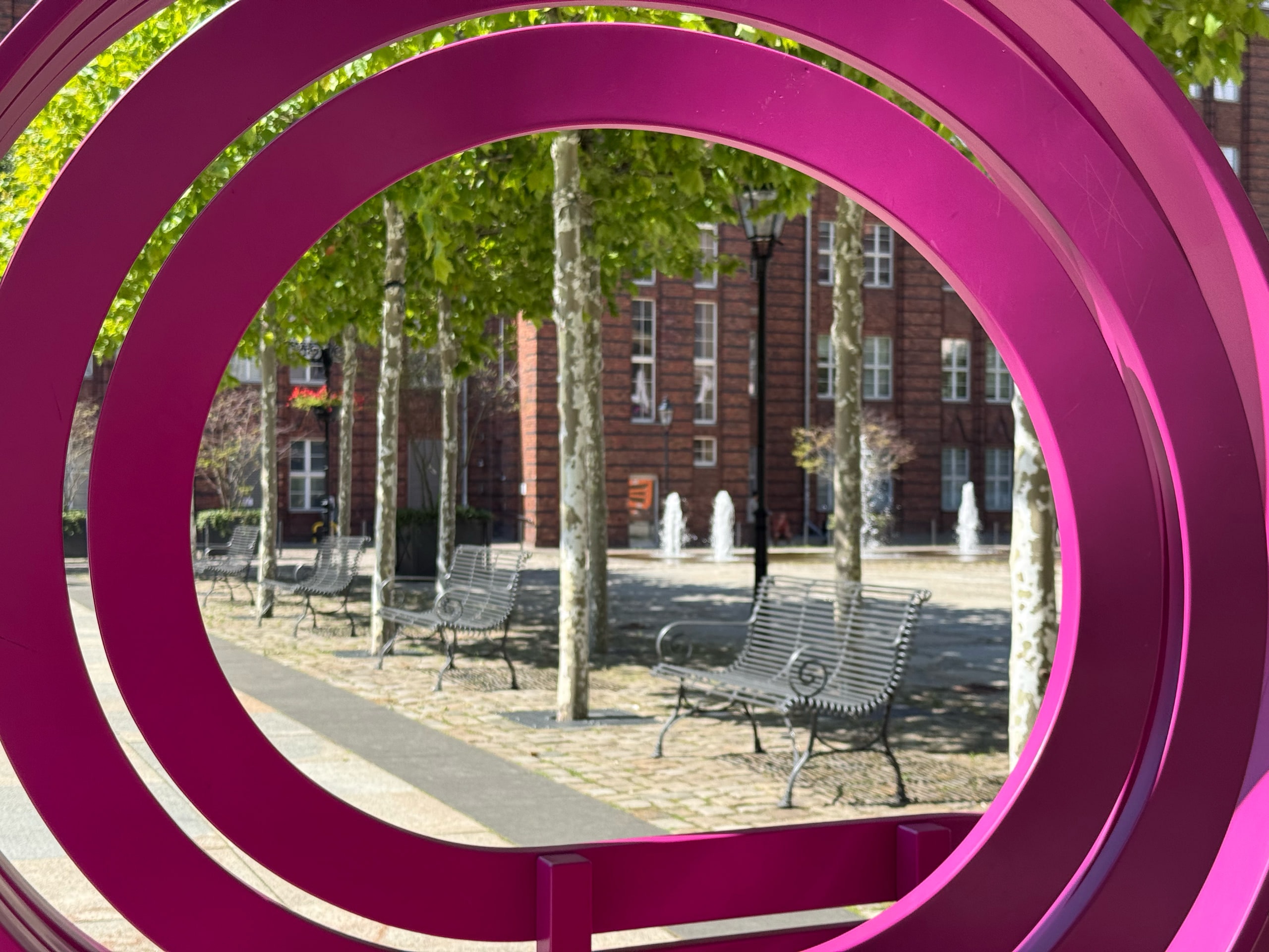 A magenta ring sculpture framing a park with fountains behind it.