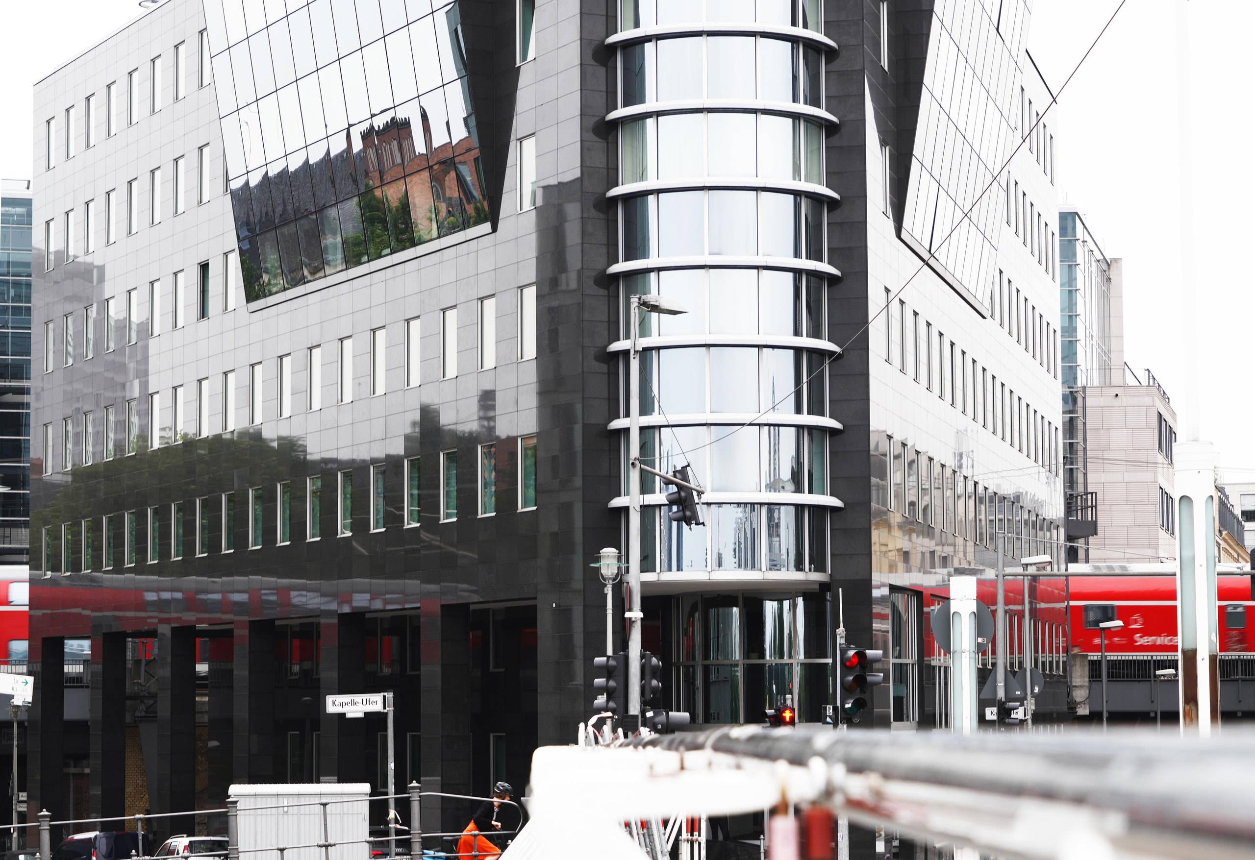 A curved glass stairwell between reflective office facades, with a red train crossing behind a bridge railing.