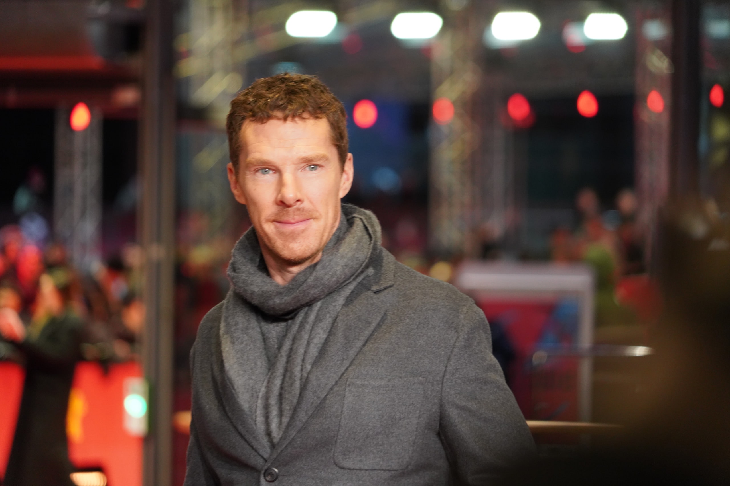 Benedict Cumberbatch in a grey coat and scarf on the red carpet at night.