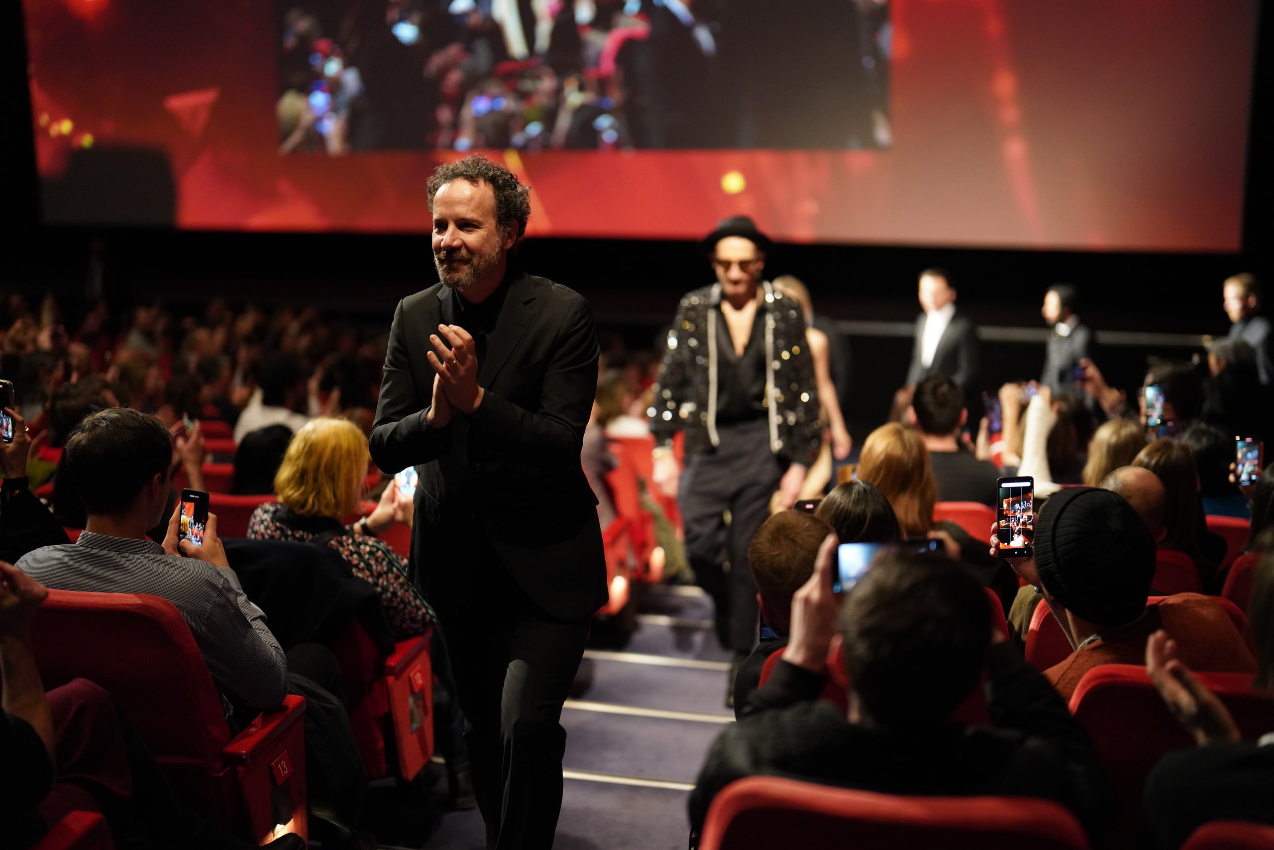 A man in a black suit applauding as he walks through a cinema aisle, surrounded by seated audience members.