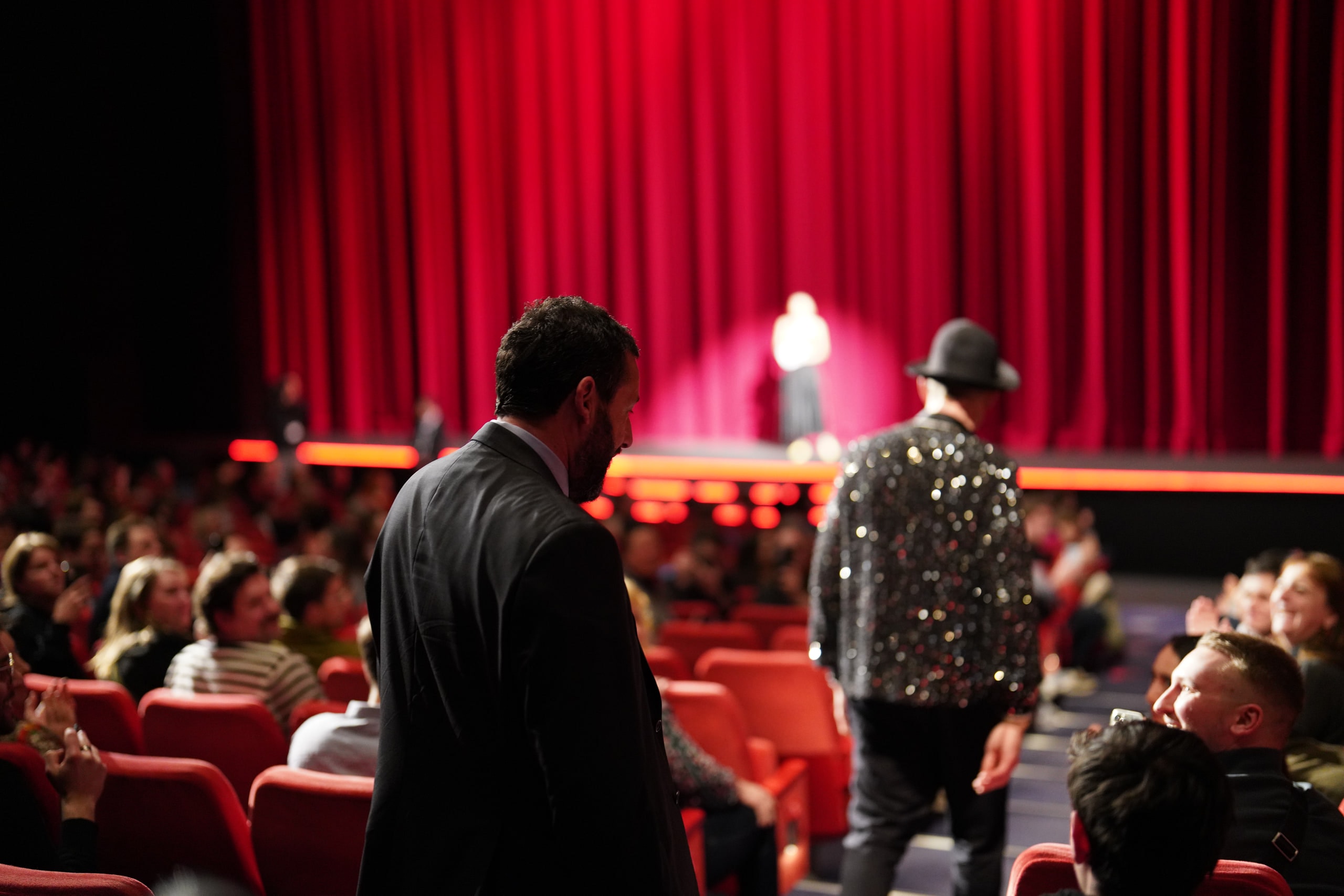 Two figures walking through the audience toward a stage framed by a red curtain, one wearing a sparkling jacket.