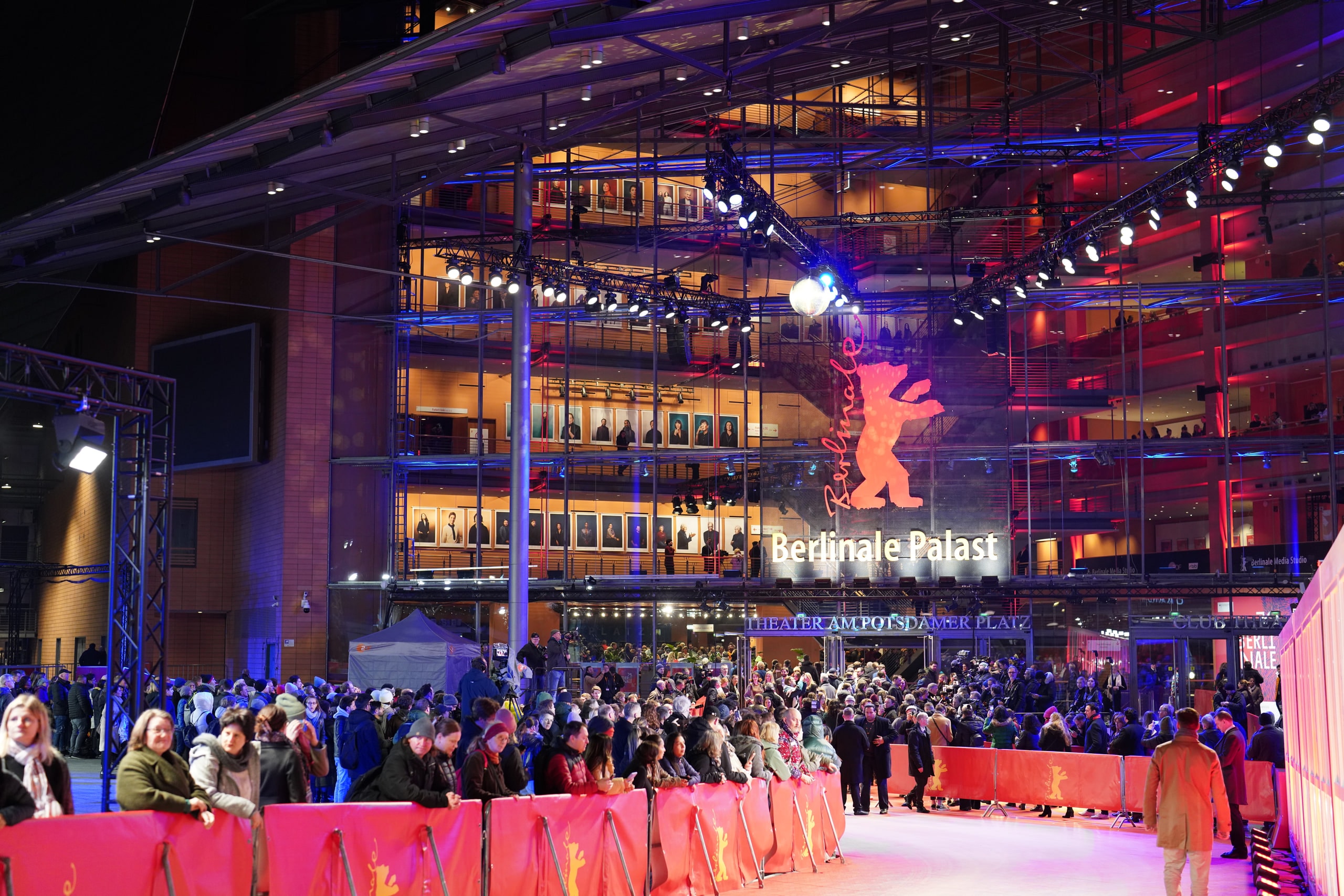 The illuminated glass facade of Berlinale Palast above a crowd gathered along red carpet barriers at night.