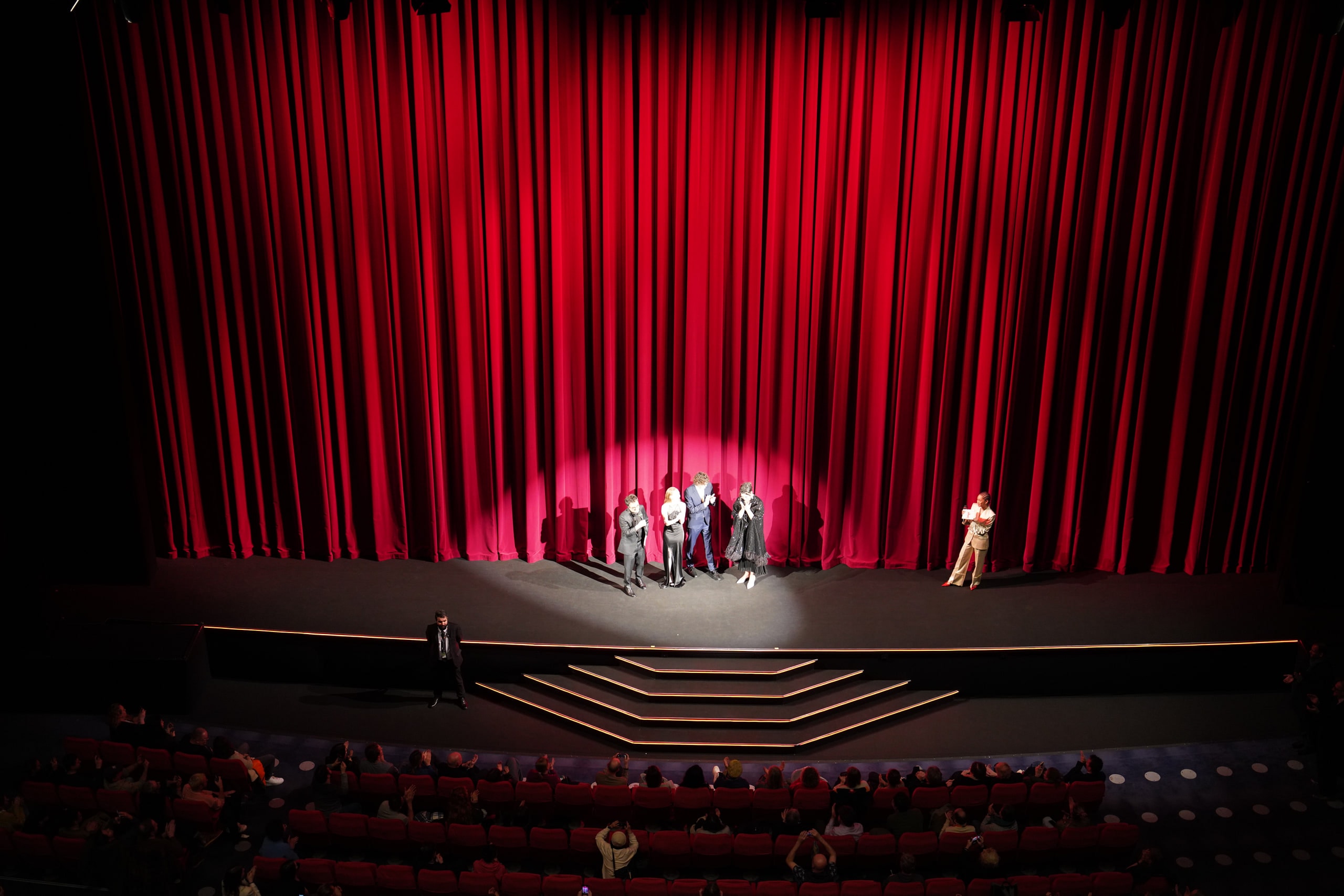 A small group on stage beneath a towering red curtain, with illuminated steps and the seated audience below.