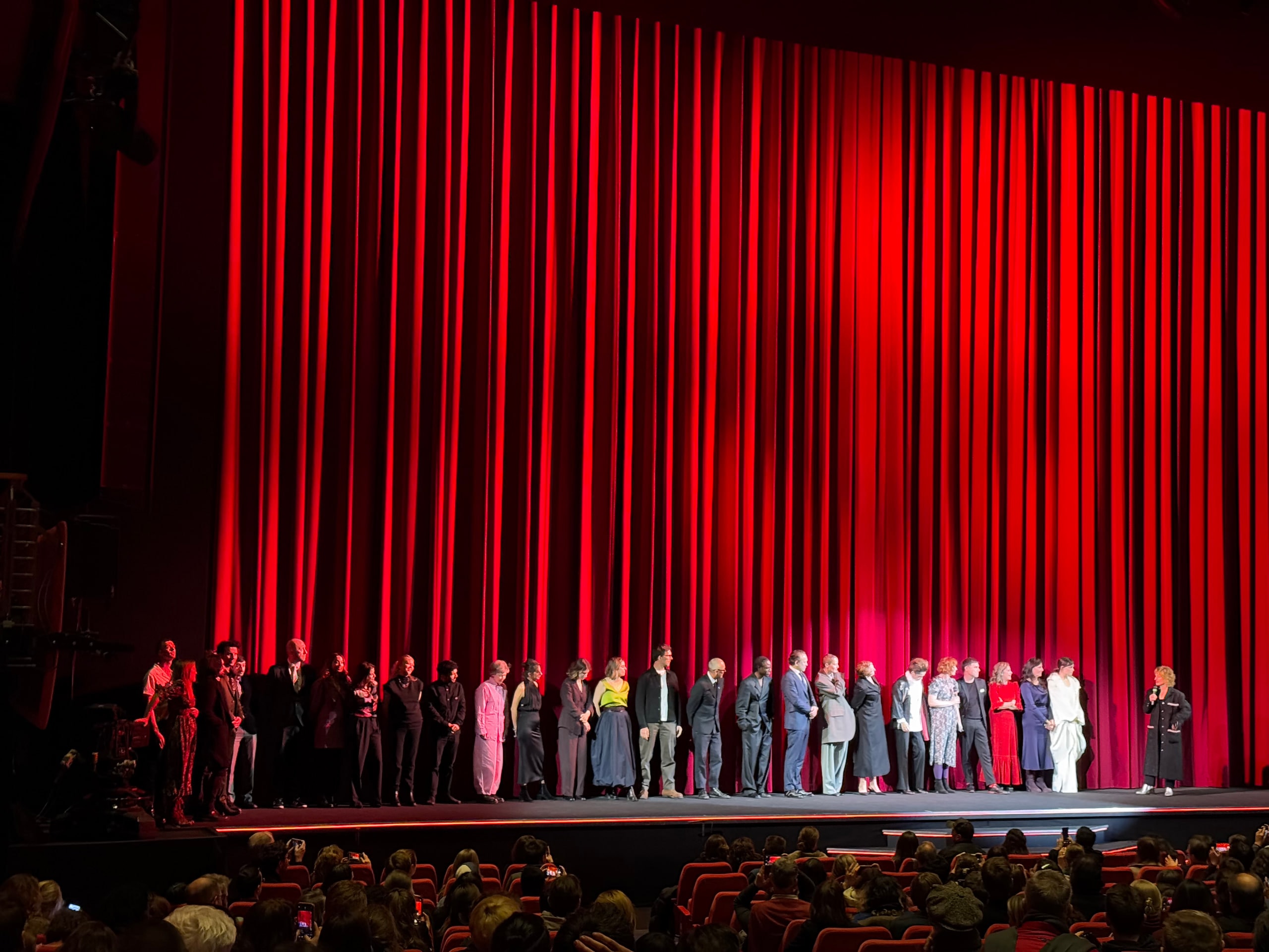 A long line of people on stage in front of red curtains, facing a speaker holding a microphone at the right.