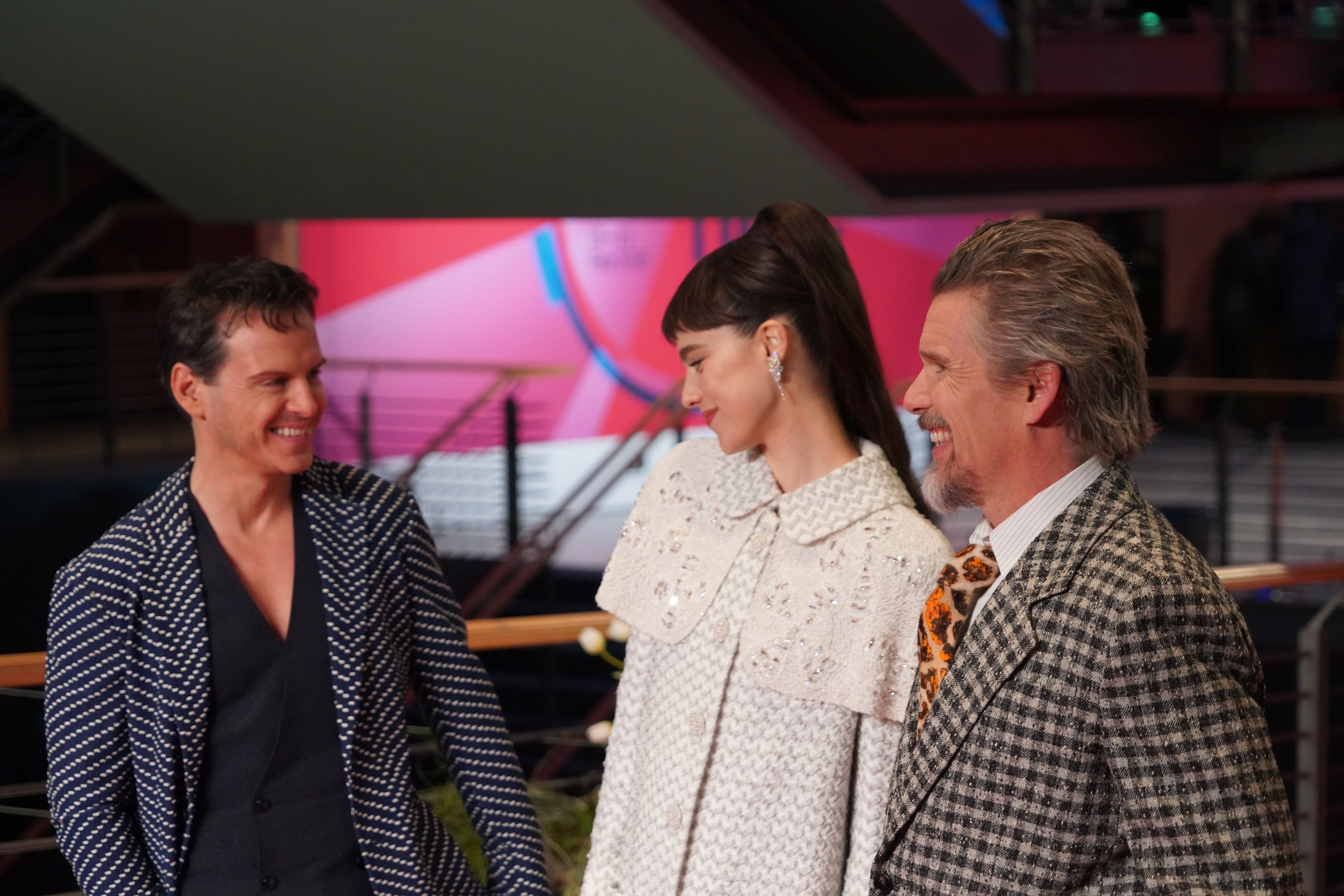 Three people sharing a smile on the red carpet, wearing a patterned dark jacket, an embellished white dress and a checked suit.