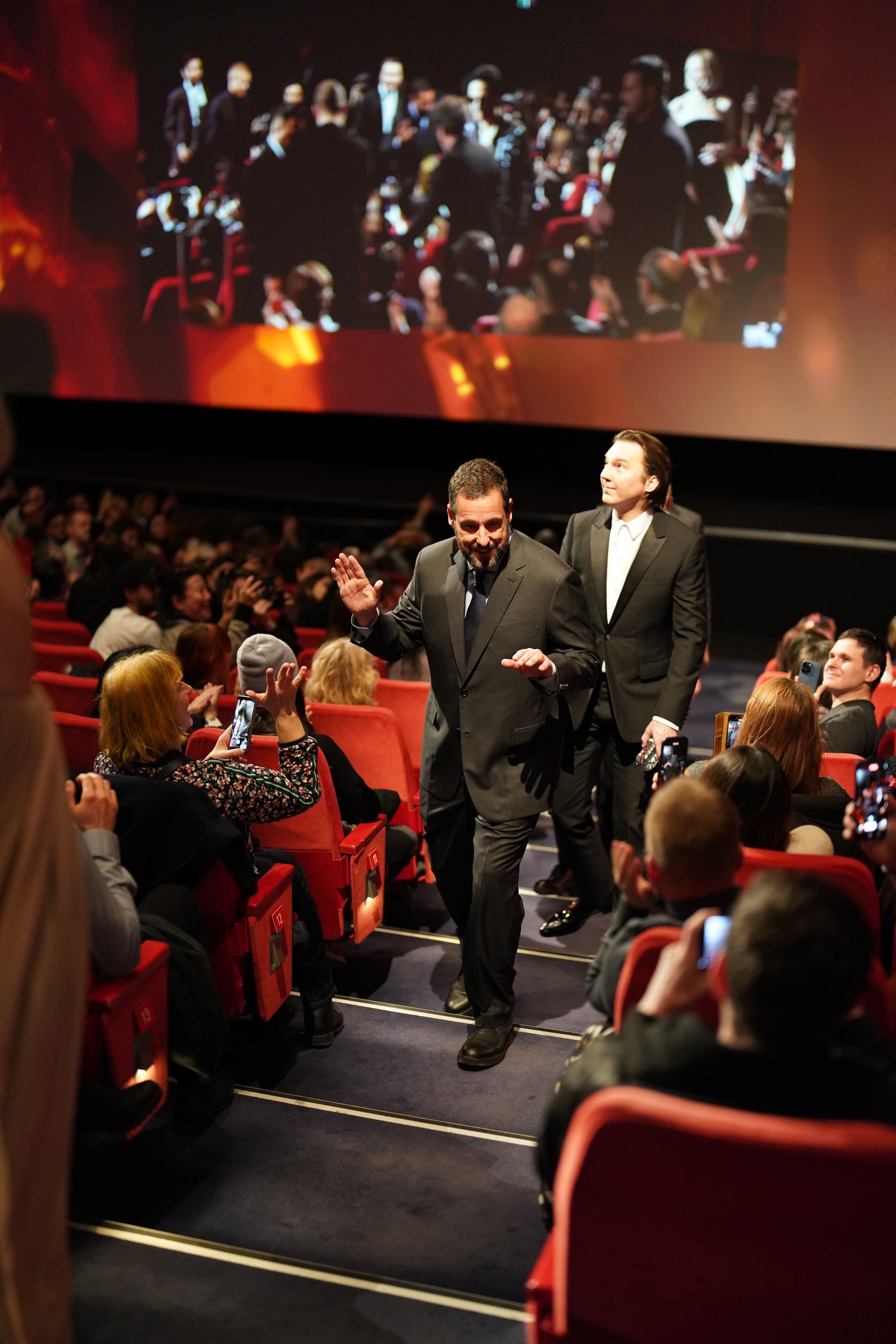 Adam Sandler raising a hand as he walks down a cinema aisle past seated audience members.