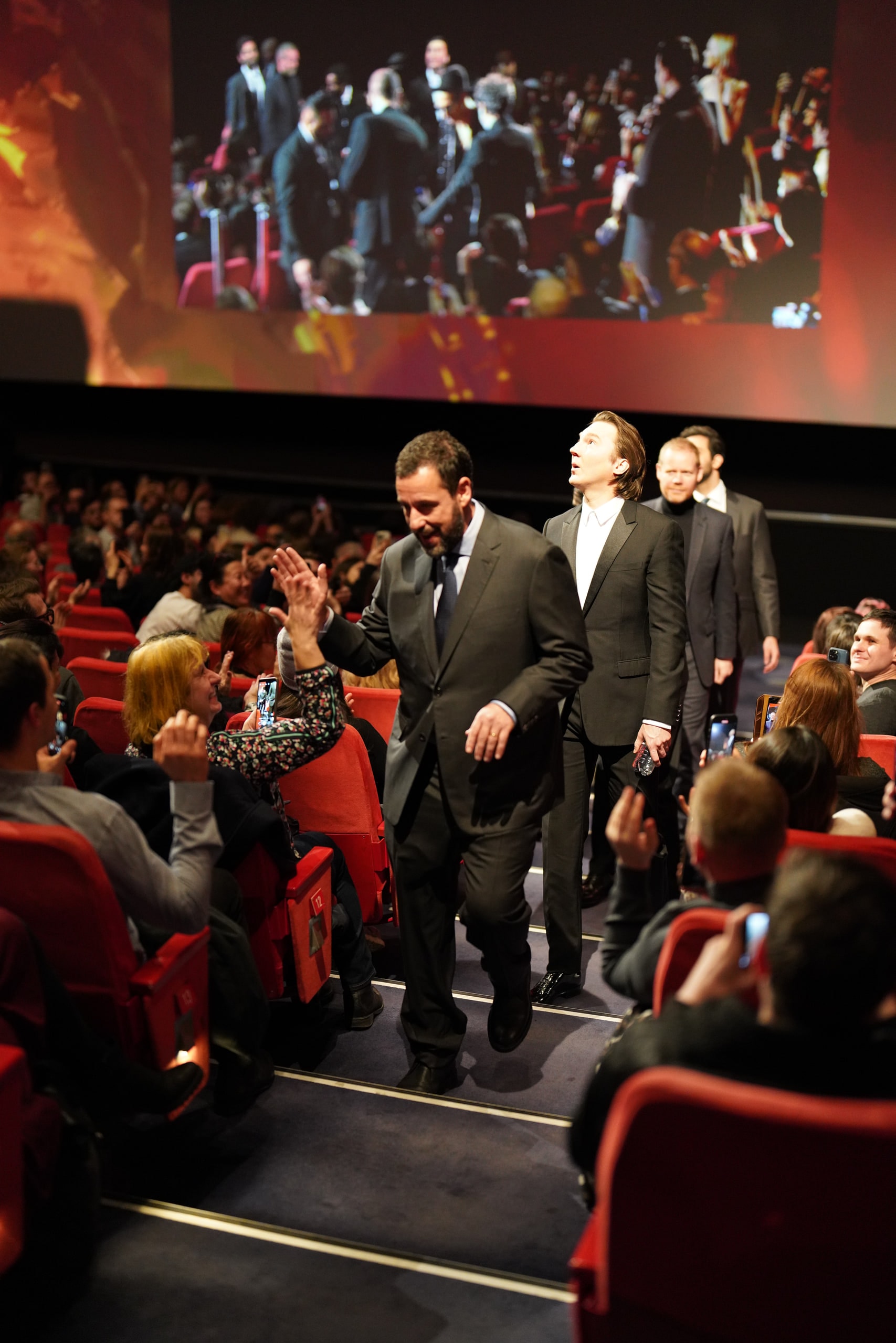 Adam Sandler greeting an audience member with a raised hand while walking through the cinema.