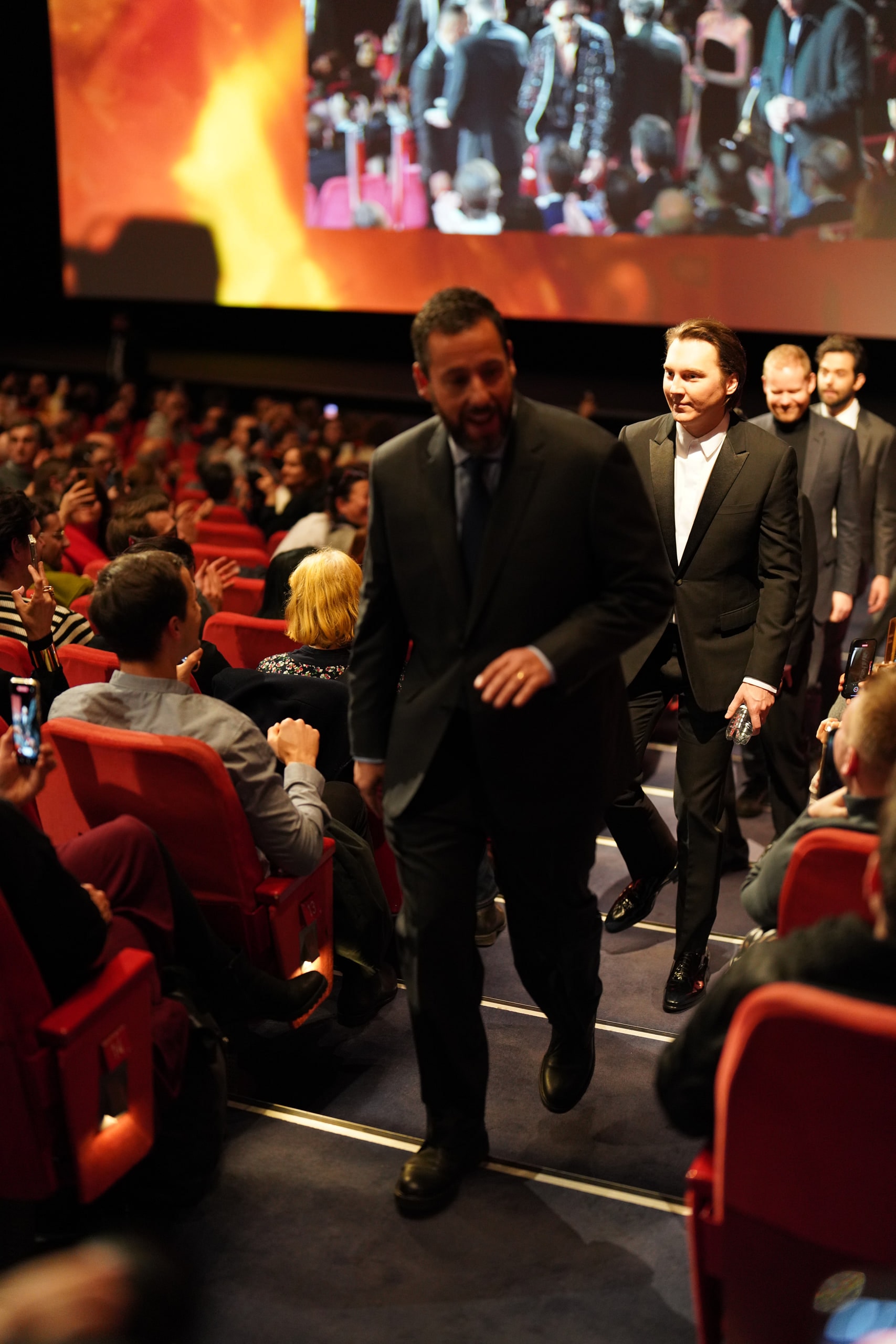 Adam Sandler approaching the camera in a dark suit, with the cinema audience on either side of the aisle.