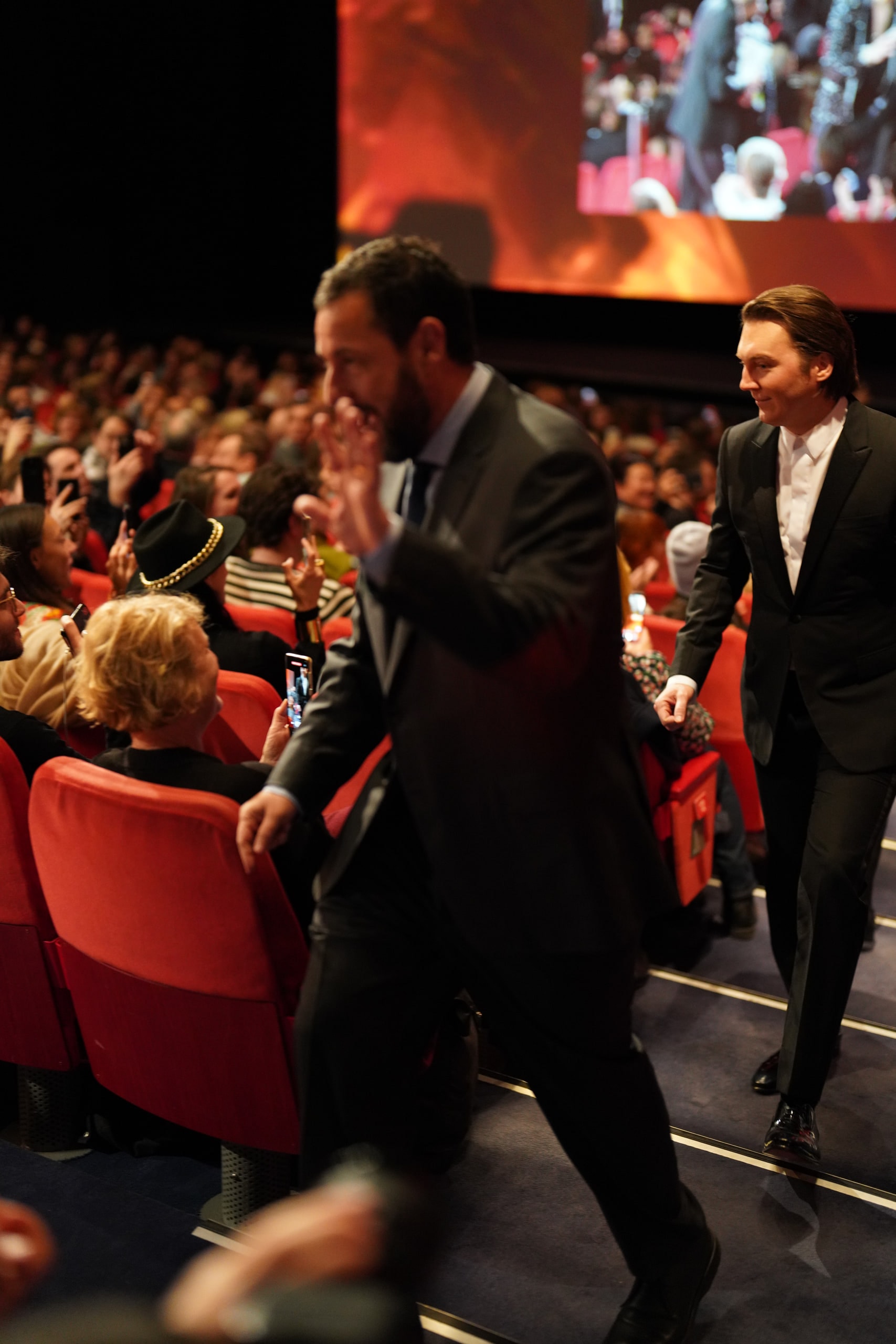 Adam Sandler passing close to the camera with a raised hand, his movement softly blurred against the cinema audience.