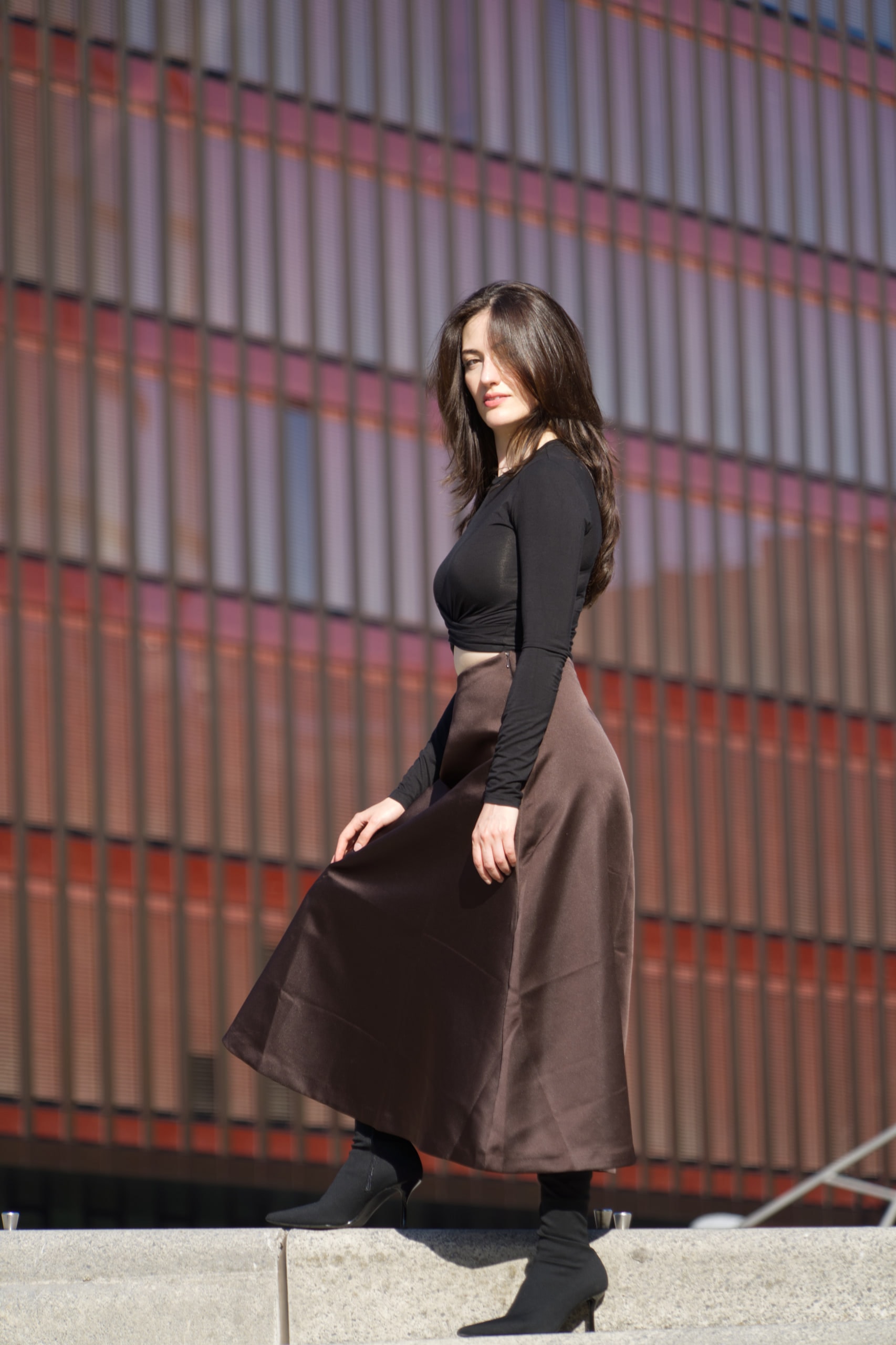 Breshna Hamid Khan turning in a brown skirt in front of a red and grey facade.