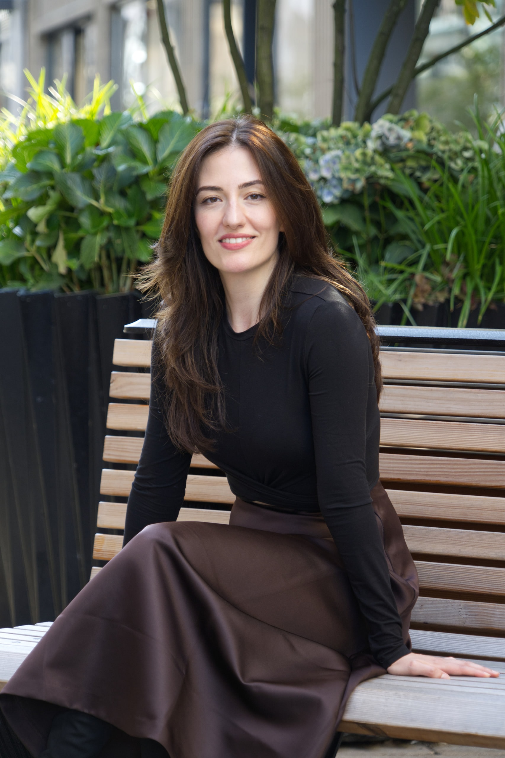 Breshna Hamid Khan smiling on a wooden bench with green plants behind her.