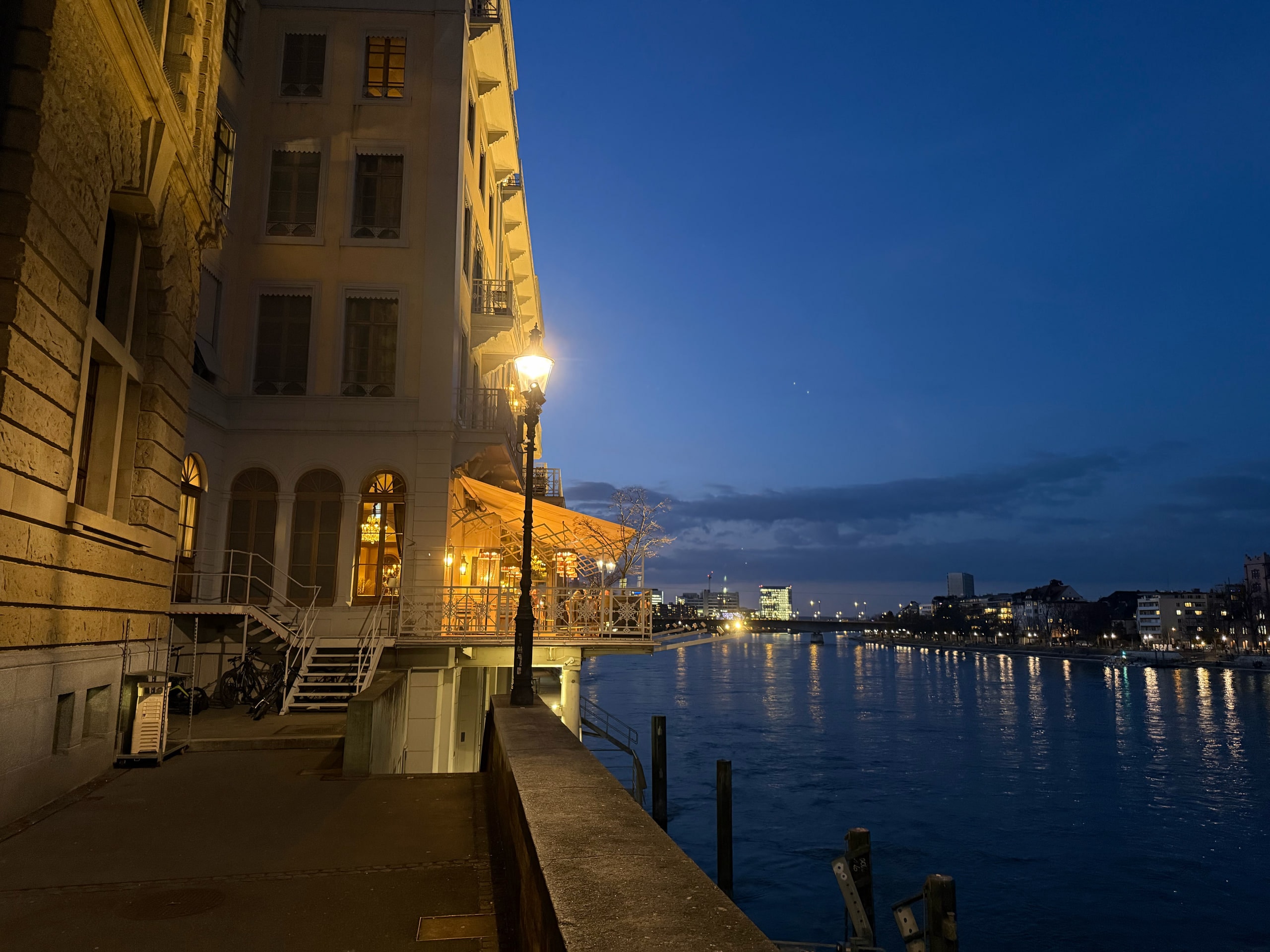 A riverside street and buildings reflected across the Rhine at blue hour.