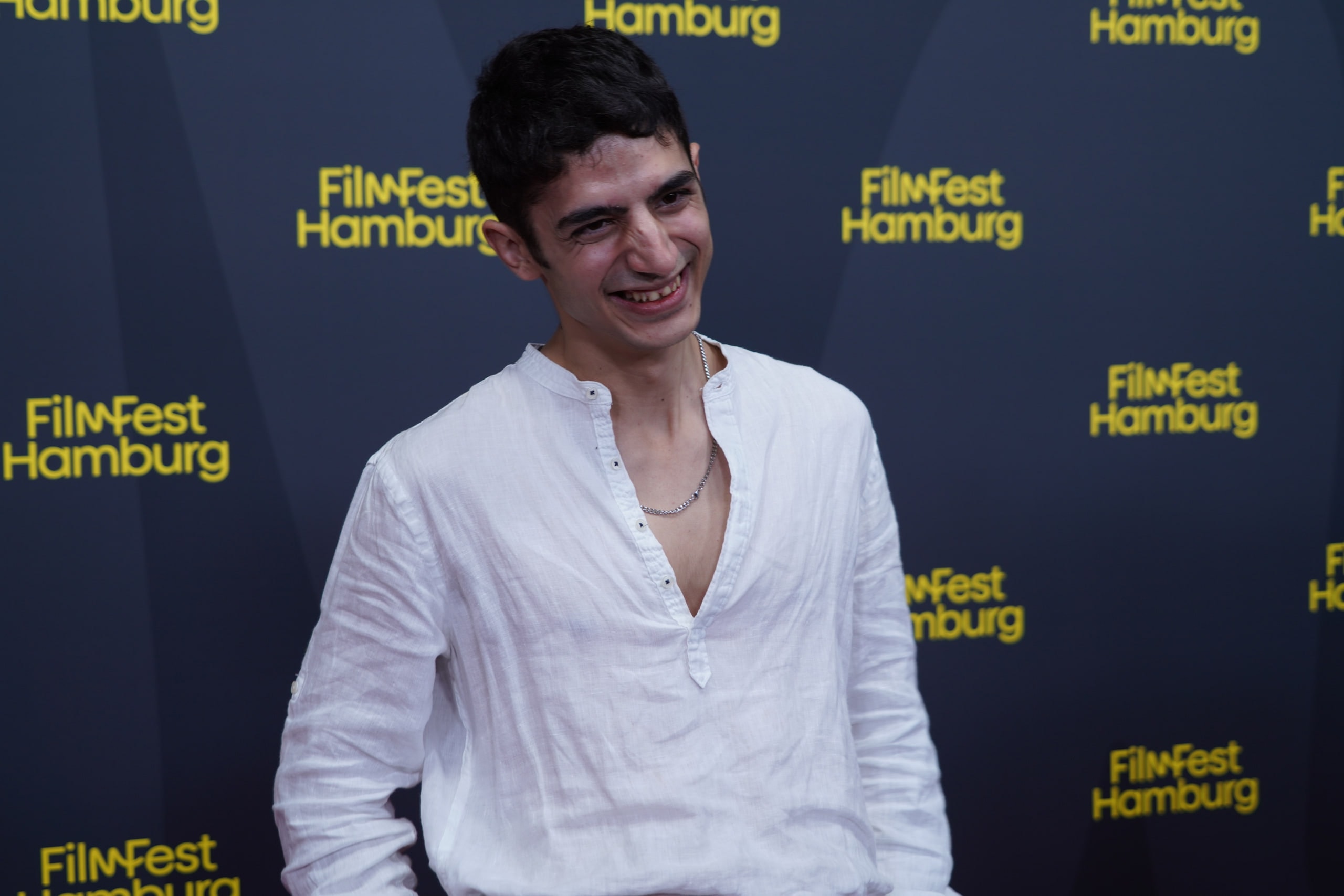 Andronikos Korakas smiling in an open-collar white shirt at the Filmfest Hamburg backdrop.