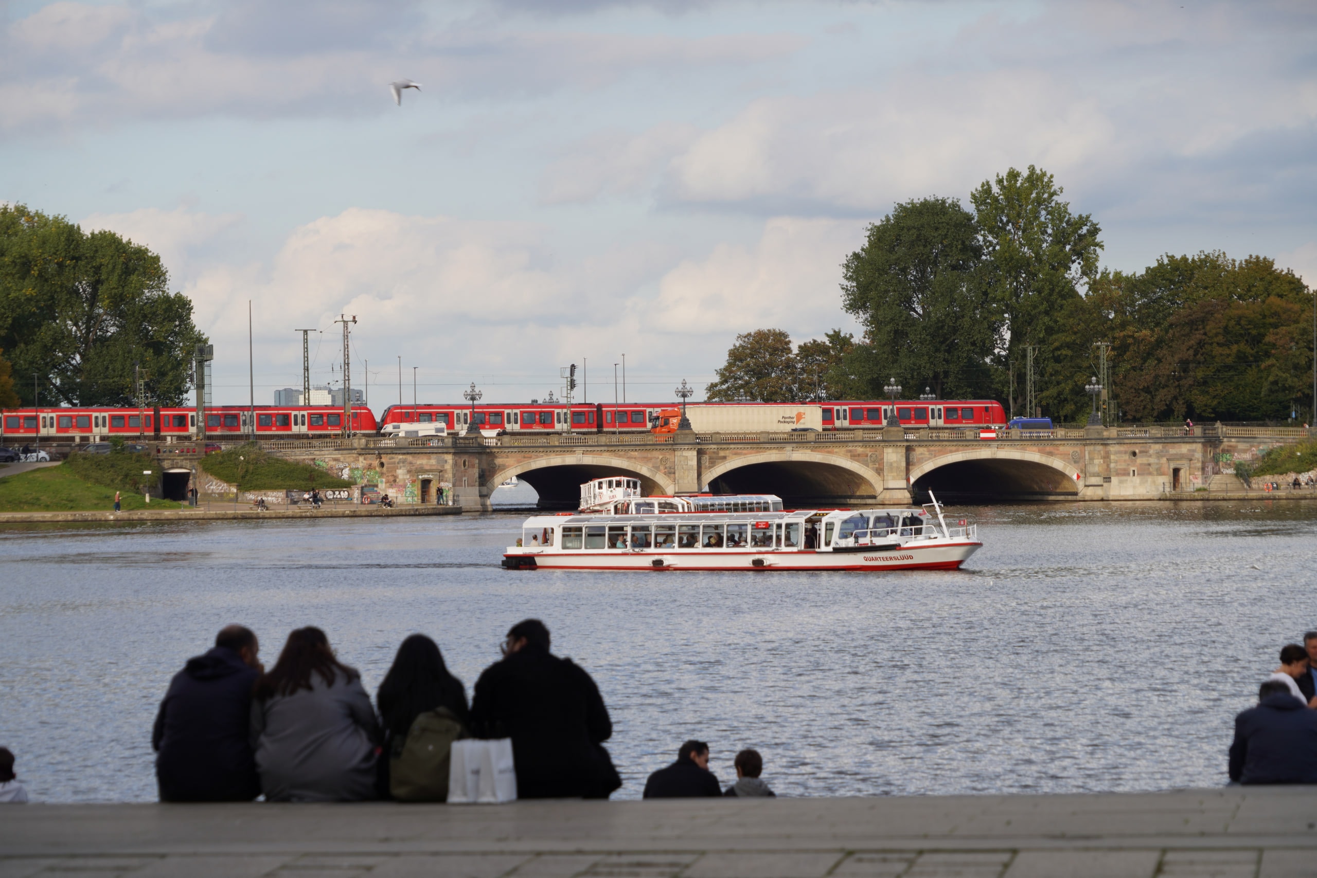 Three people sitting beside the water in Hamburg as a sightseeing boat passes beneath a bridge.