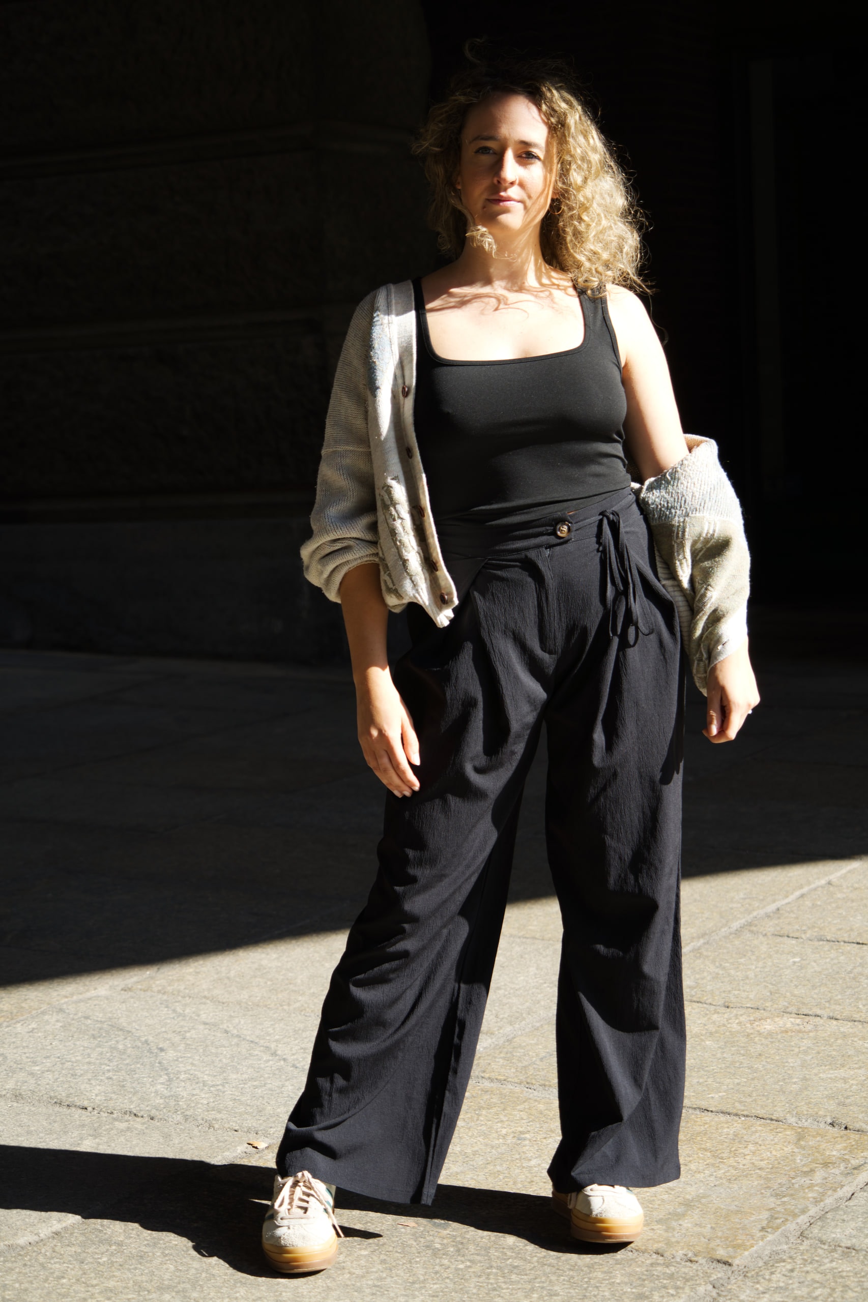 Larissa Kählert standing at the edge of sunlight and deep shade, wearing black trousers and a grey cardigan.