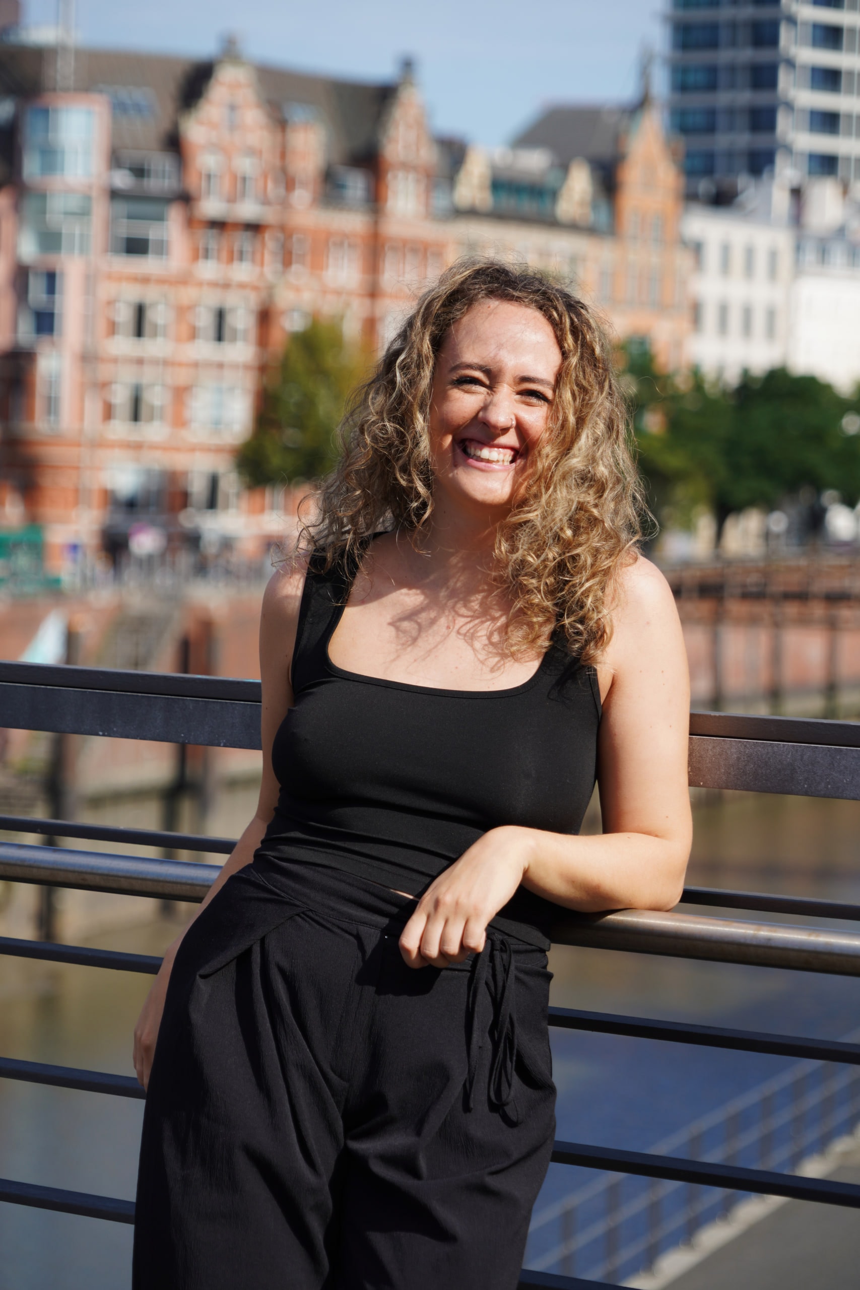 Larissa Kählert laughing beside a black waterside railing, Hamburg's brick buildings behind her.