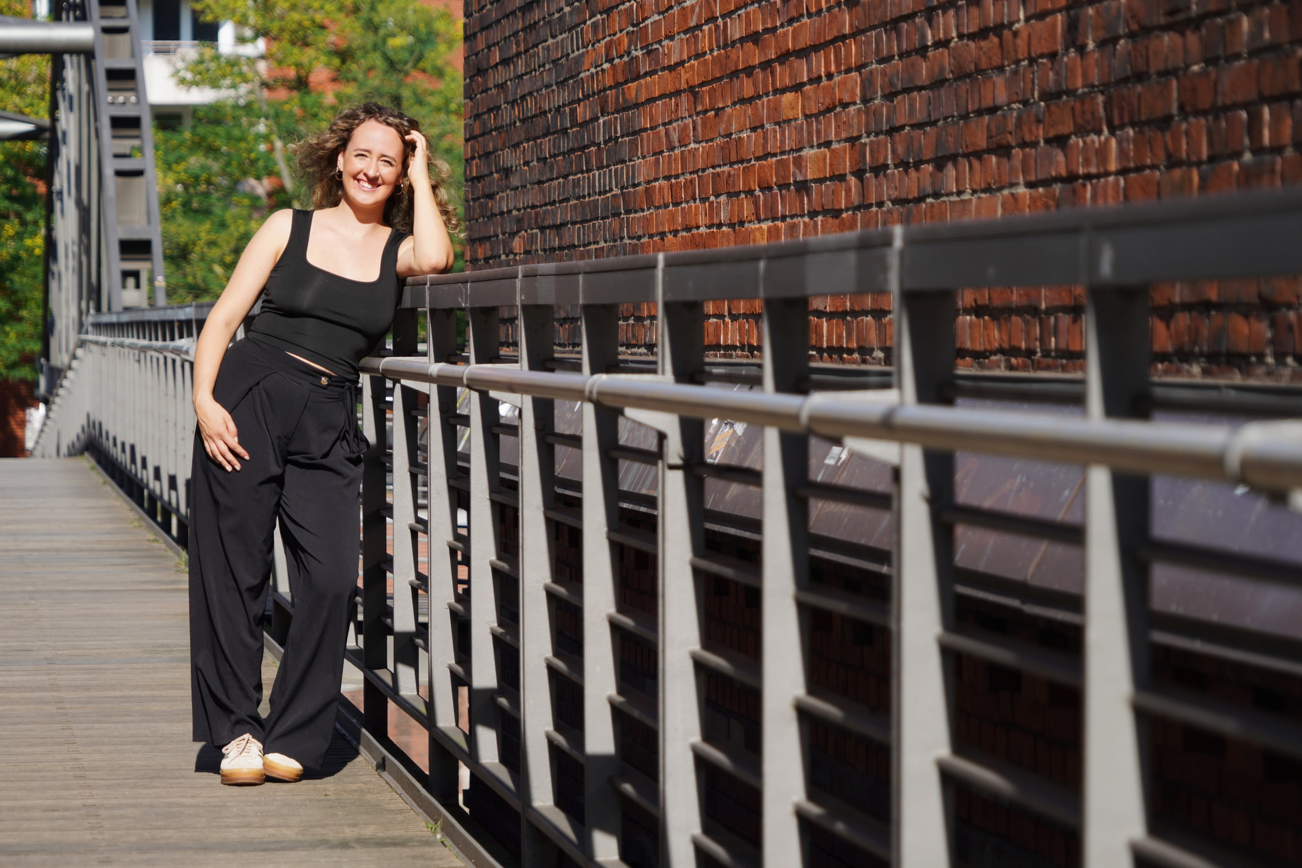 Larissa Kählert smiling on a wooden footbridge, one hand raised to her hair beside a brick wall.