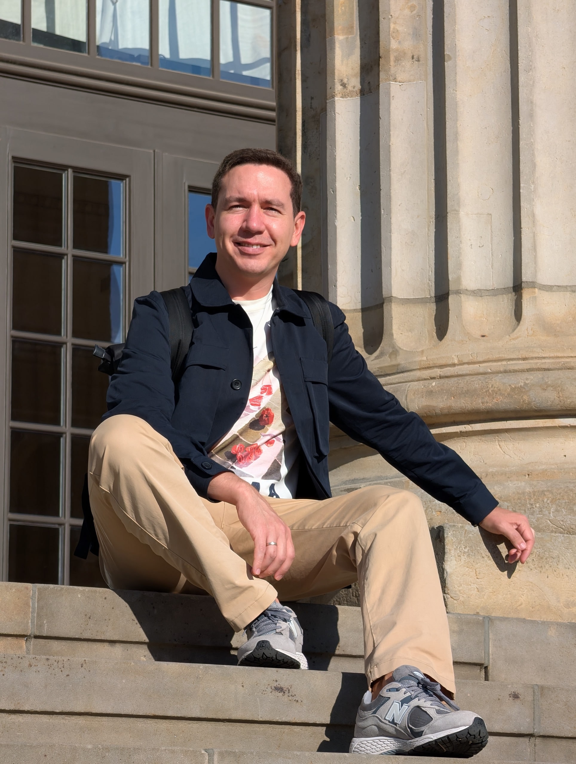 Ramis Vakazov seated on stone steps beside a column at Gendarmenmarkt in Berlin, wearing a navy jacket and beige trousers in sunlight.