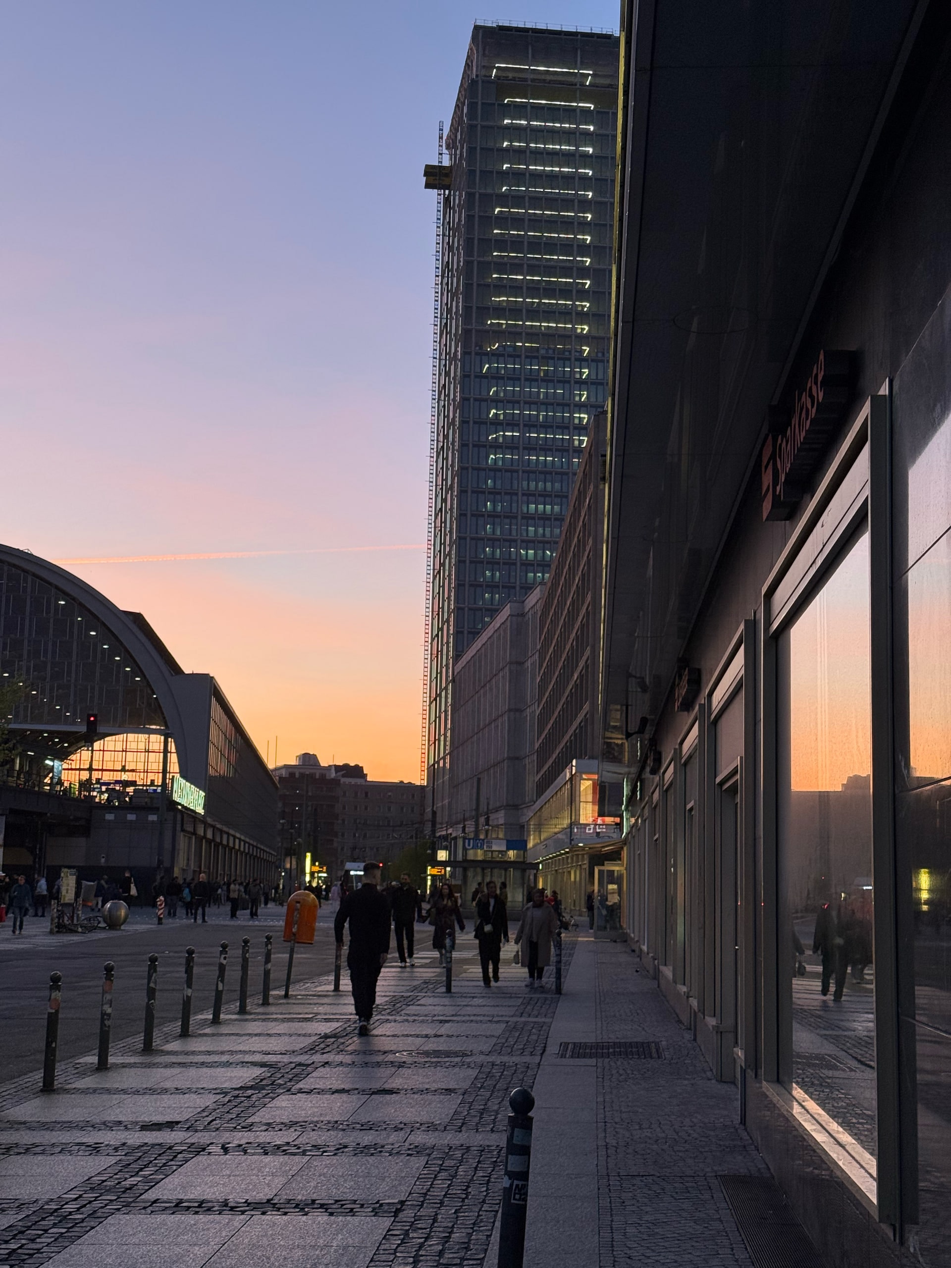 People walking a wide street at dusk beneath a dark tower.