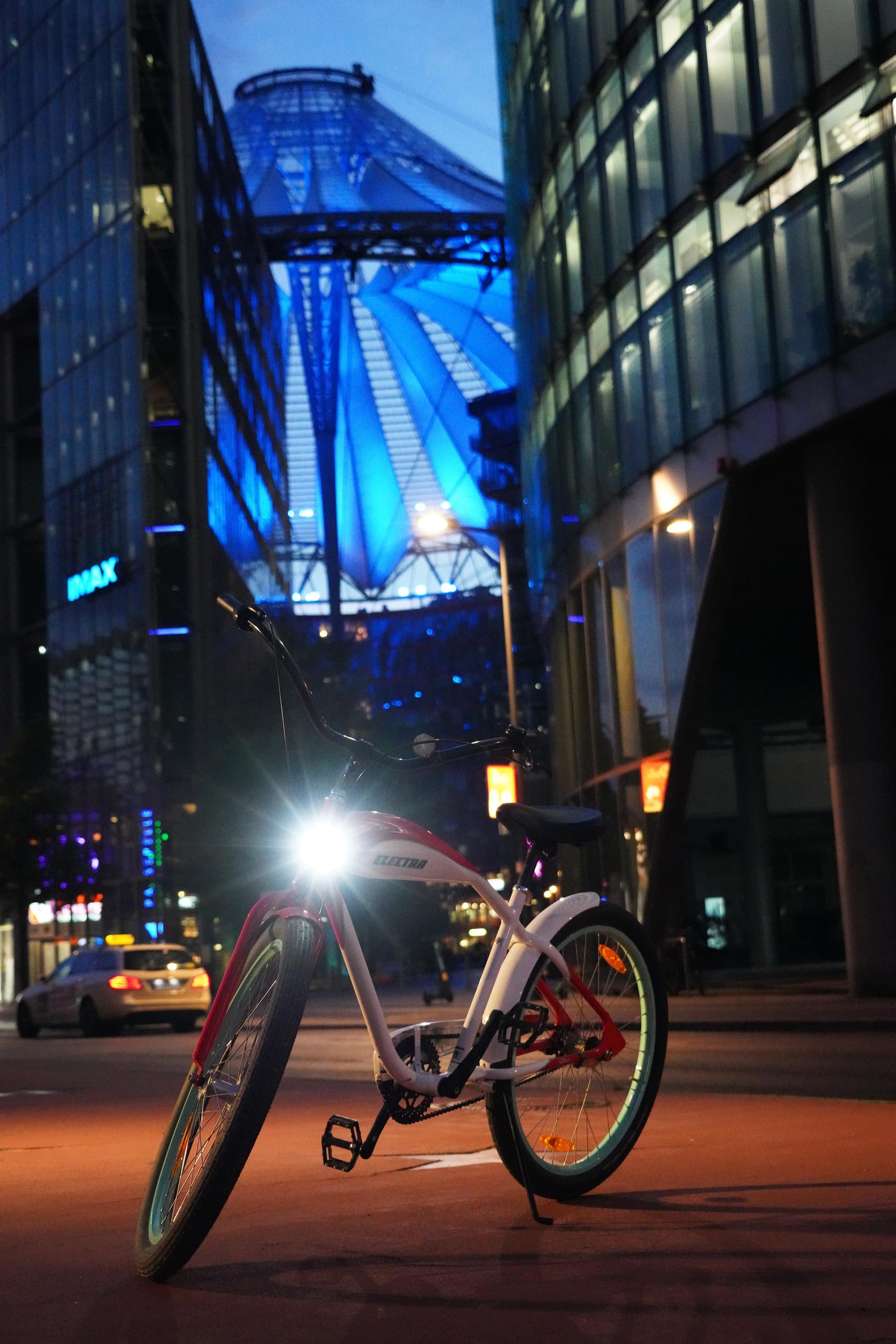 A bicycle with a bright headlight on a quiet street at night, framed by glass buildings and a blue illuminated canopy.