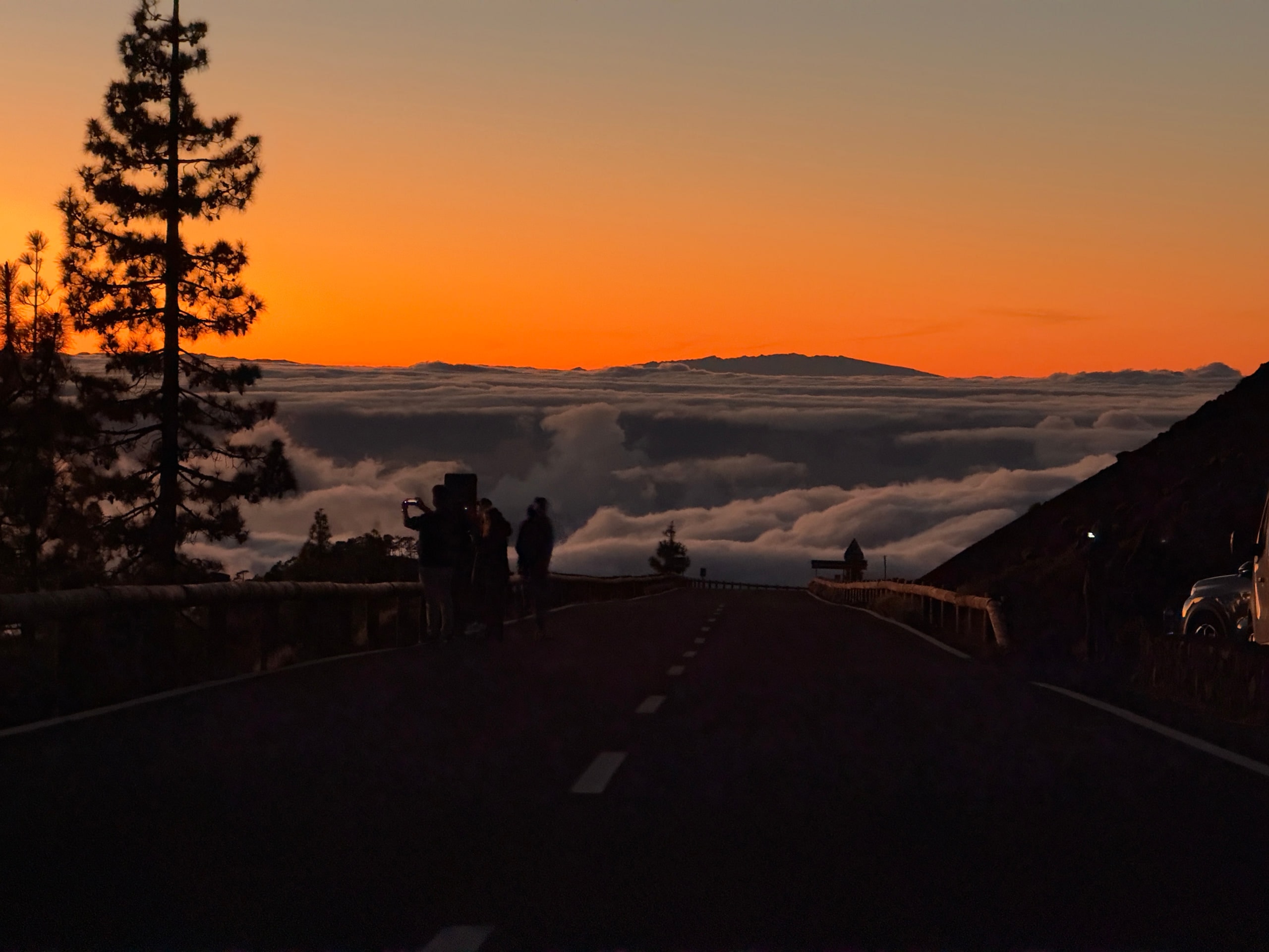 A mountain road descending toward a sea of clouds beneath an orange sunset.