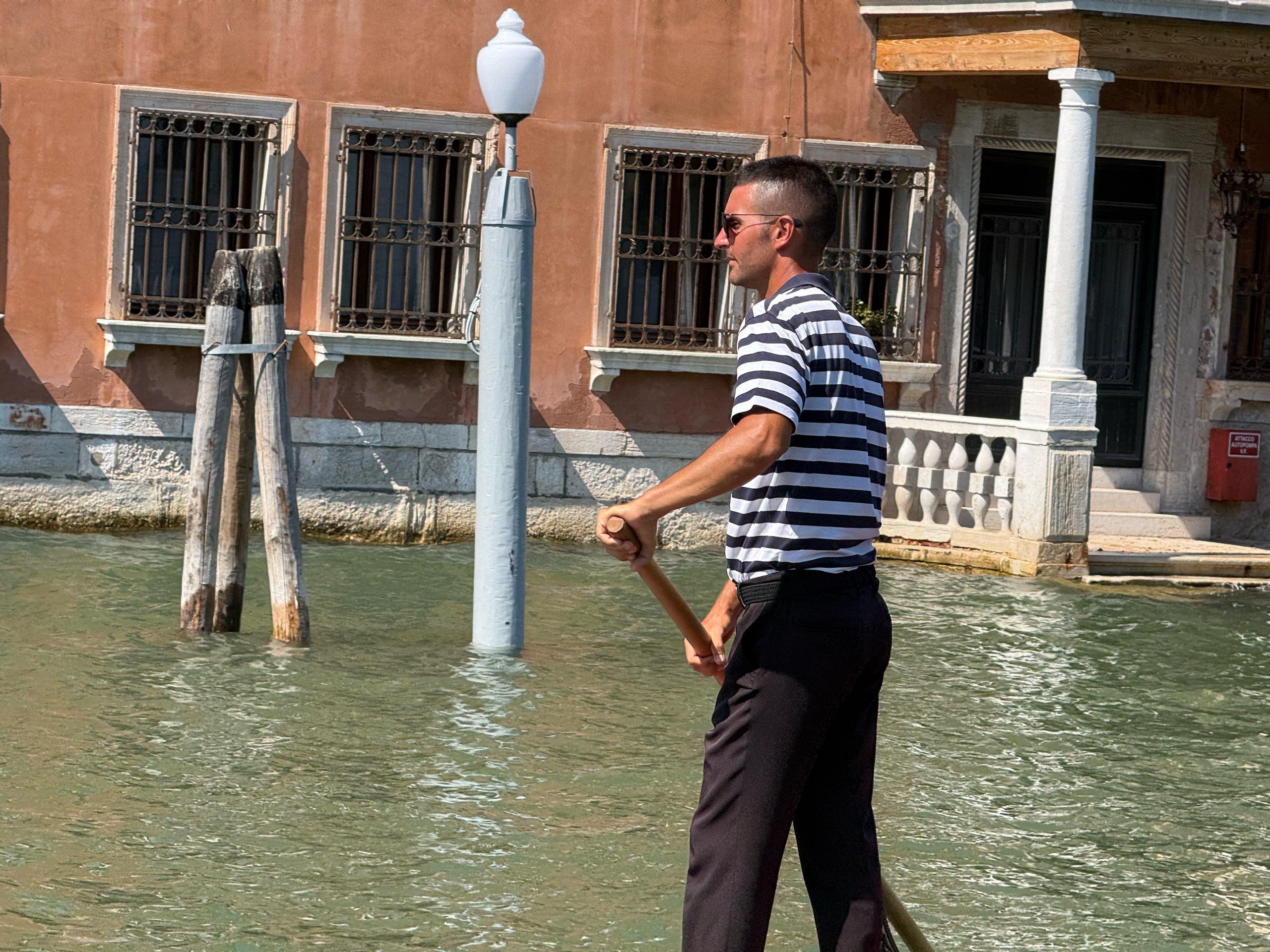 A gondolier in a striped shirt resting on his oar beside the mooring poles.