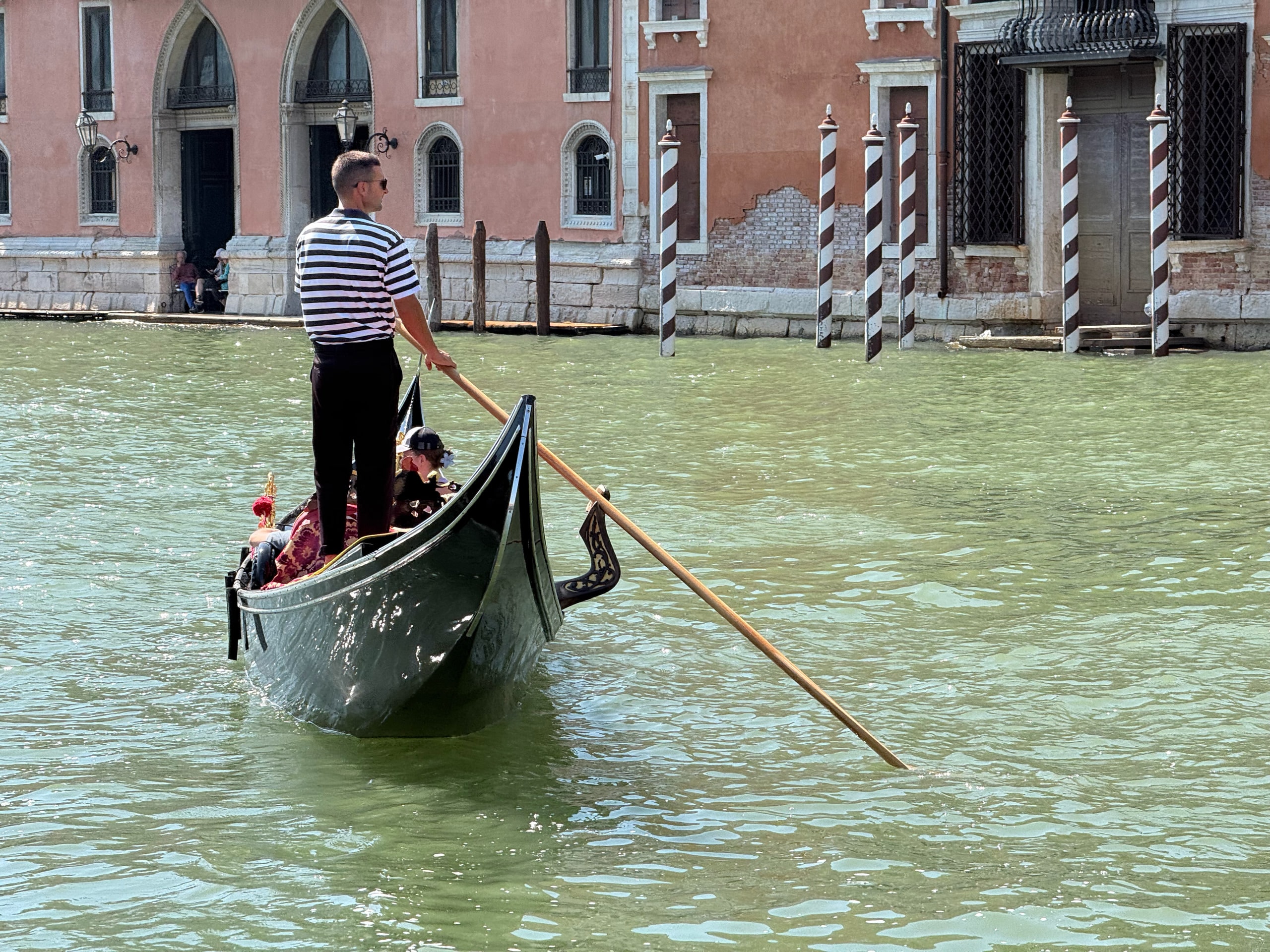 A gondolier steering across green water past a brick palazzo.