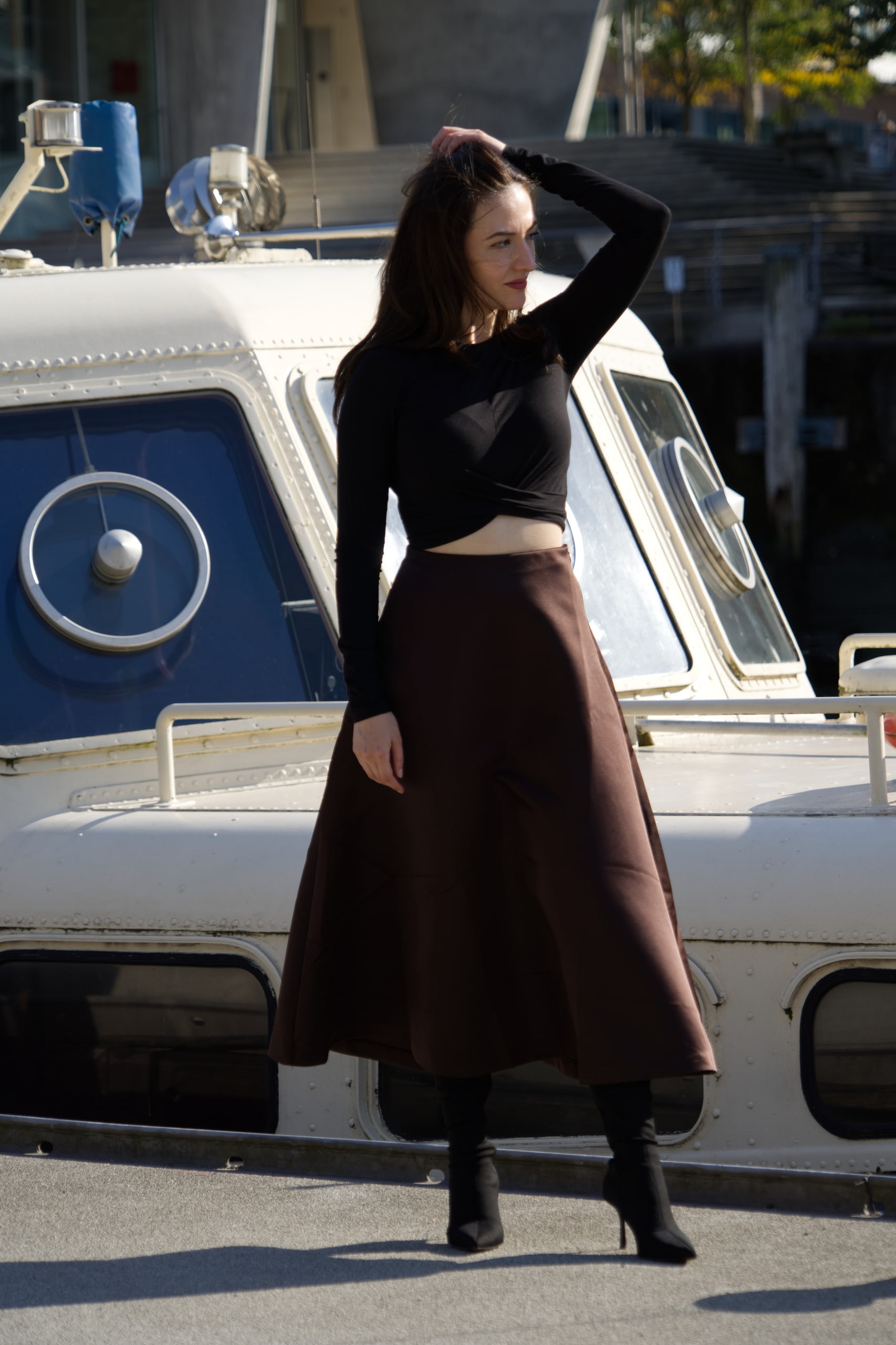 Breshna Hamid Khan in a black top and brown skirt beside a white boat on Hamburg's waterfront.