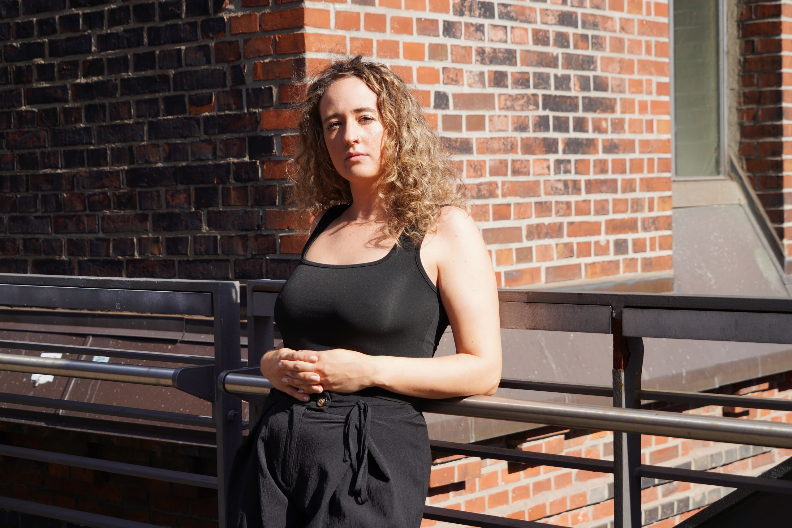 Larissa Kählert standing beside a railing where sunlight meets shadow on a brick wall.