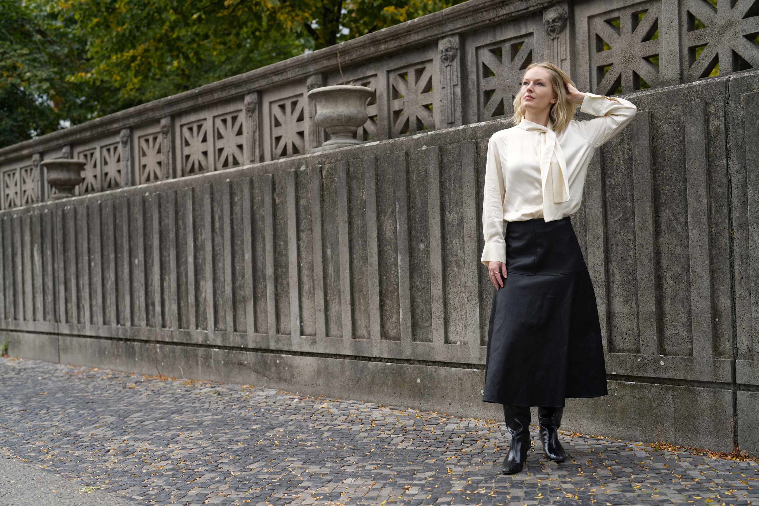 Sophia Repp beside a long stone balustrade, one hand raised to her hair beneath autumn trees.