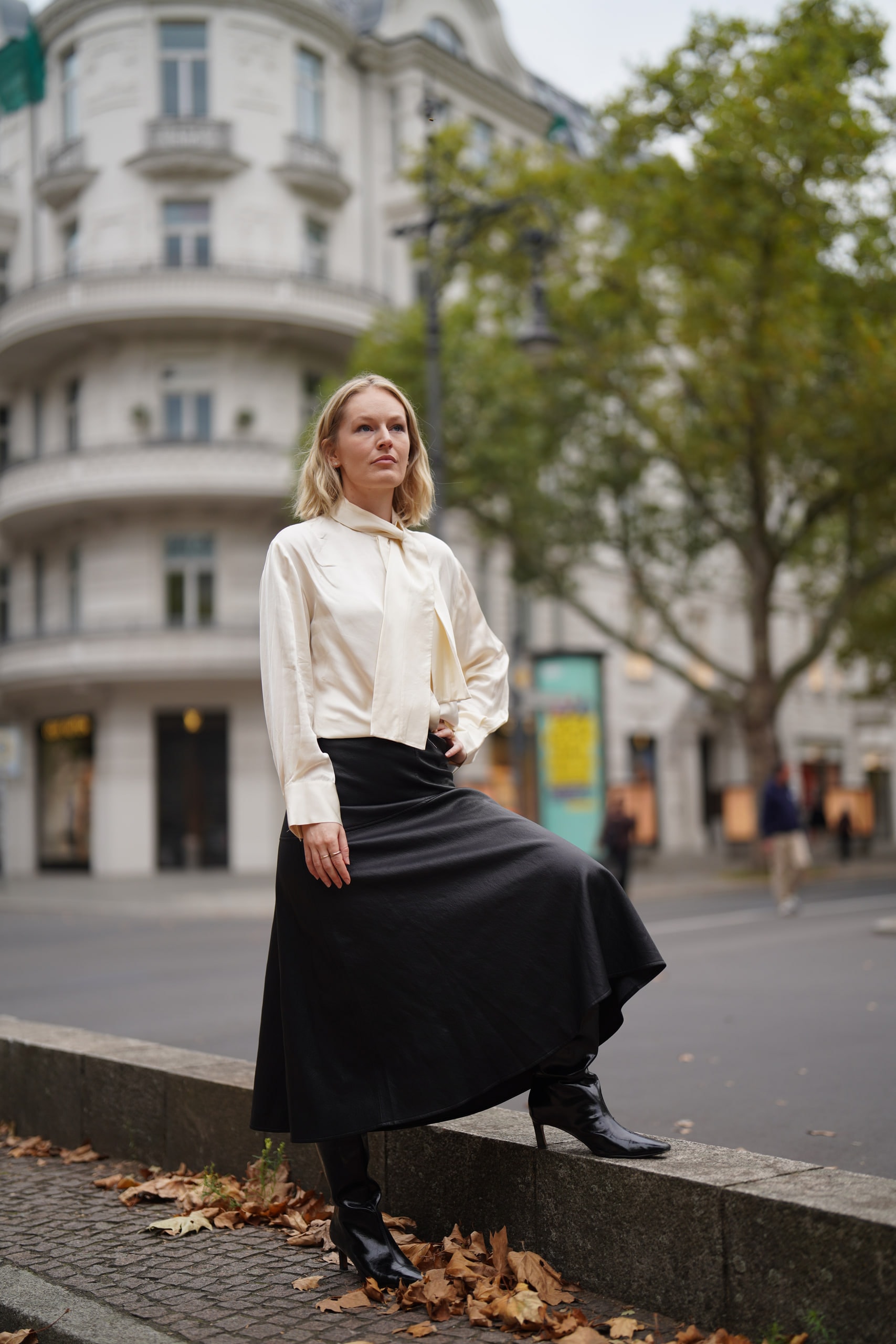 Sophia Repp resting one boot on a low stone wall at a Berlin street corner, her black skirt spread over her knee.
